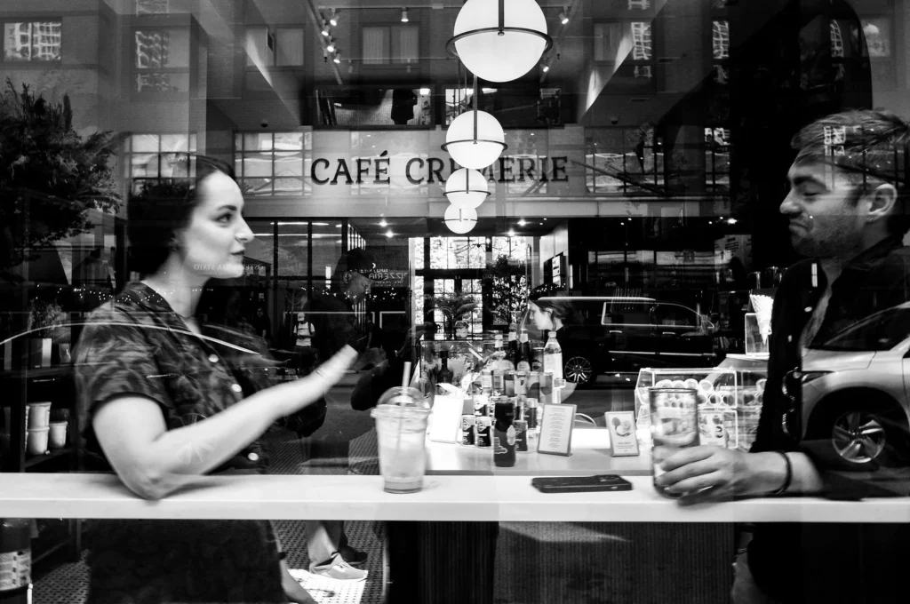 Black and White photo of couple eating at a table near a window. The perspective is of someone looking into the restaurant.