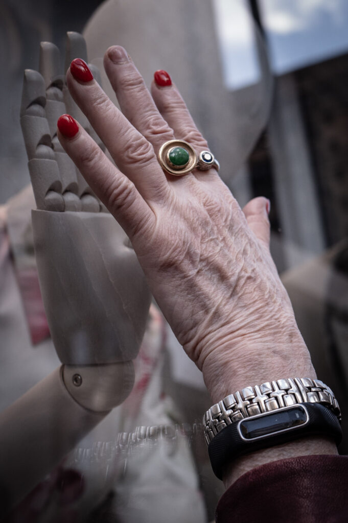 Hand touching a mannequin's hand through a window pane.
